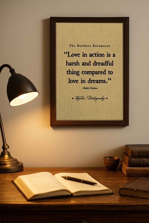 Framed “Love in Action” quote from The Brothers Karamazov by Dostoyevsky displayed above a wooden writing desk with an open book and brass lamp casting a soft glow.