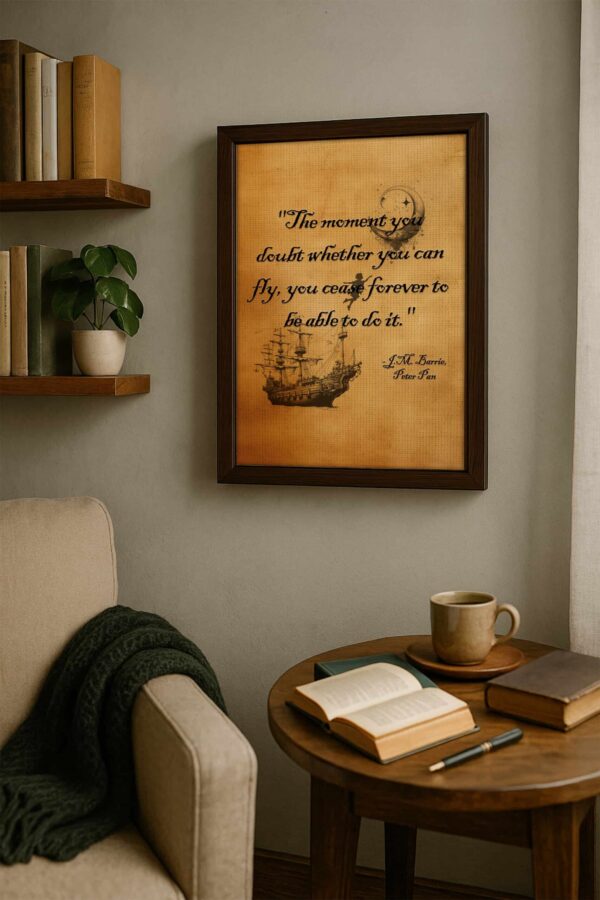 J.M. Barrie quote print in a dark wood frame hanging on a gray wall above a cozy reading nook with books, a green throw, and a coffee mug.