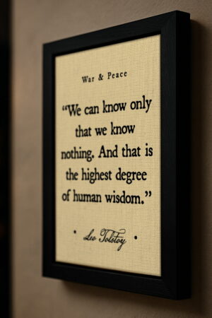 Side angle of the Tolstoy quote in a black frame mounted on a warm taupe wall, showing canvas texture and frame depth with soft directional lighting.